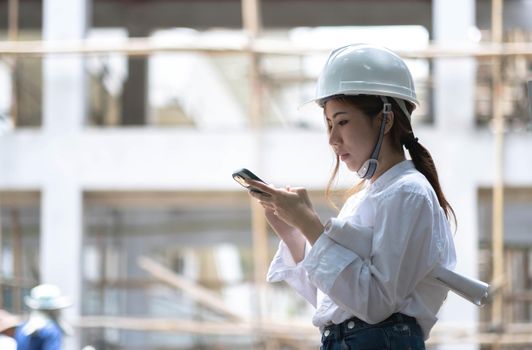 Architect with a blueprints at a construction site. Portrait of woman constructor wearing white helmet and safety yellow vest. Upset sad, skeptical, serious woman looking at the phone screen outdoors..