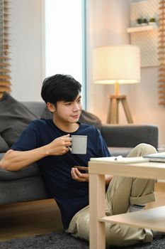 Relaxed asian man sitting on carpet in living room and reading online news on laptop.