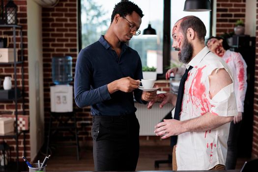 African american adult chatting with zombie in business office, undead spooky corpse chatting with person in startup workplace. Evil horror macabre monster with bloody scars and decayed.