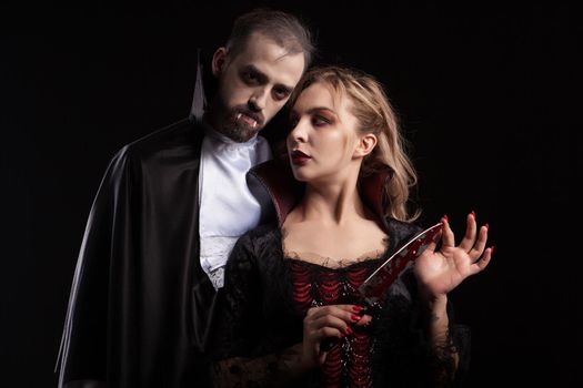 Beautiful young vampire woman with a blade covered in blood looking at her man dressed up like Dracula for halloween. Seductive couple.