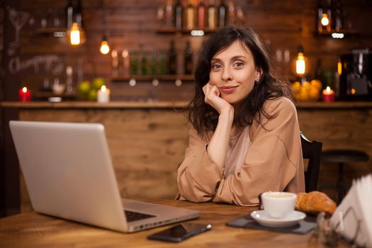 Portrait of cheerful young woman sitting down in a coffee shop smilling at the camera. Beautiful woman in a coffee shop with modern computer. Coffee shop with internet connection.