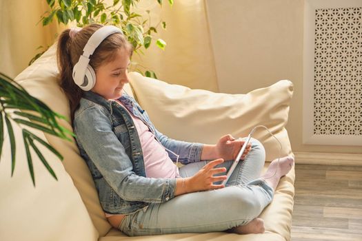 Cute little girl in casual clothes and headphones using a tablet and listening to music while sitting on a sofa in the room.
