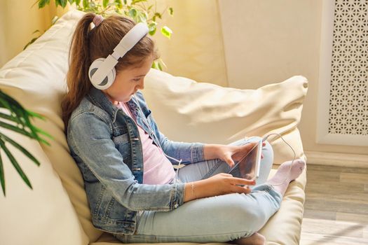 Cute little girl in casual clothes and headphones using a tablet and listening to music while sitting on a sofa in the room.