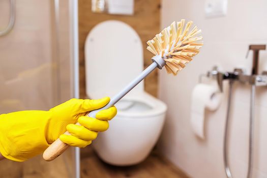 close-up, a hand in a yellow rubber cleaning glove holds a toilet brush for sanitary cleaning of the toilet. Modern bathroom background, copy space and your design. Beautiful interior
