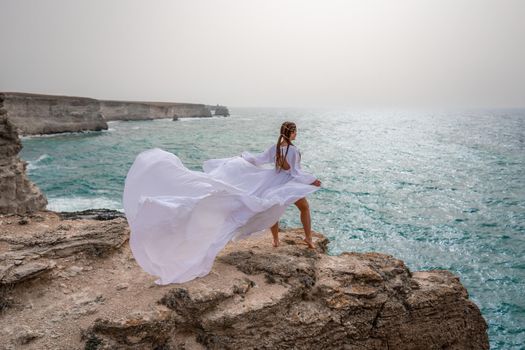 Happy freedom woman on the beach enjoying and posing in white dress over the sea. View of a girl in a fluttering white dress in the wind. Holidays, holidays at sea