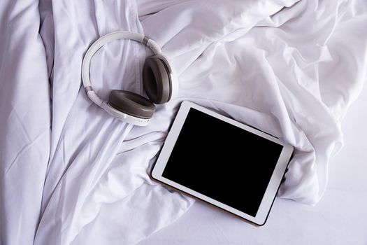 Top view, close-up. Crumpled white bedding early in the morning. There are headphones and a tablet on the bed. Mockup for tablet, black screen. Modern technologies.