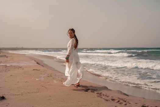 Model in boho style in a white long dress and silver jewelry on the beach. Her hair is braided, and there are many bracelets on her arms