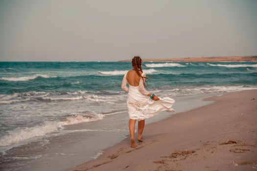 Model in boho style in a white long dress and silver jewelry on the beach. Her hair is braided, and there are many bracelets on her arms