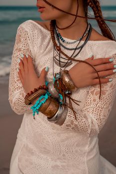 Model in boho style in a white long dress and silver jewelry on the beach. Her hair is braided, and there are many bracelets on her arms