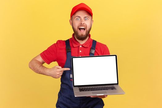 Portrait of excited bearded handyman in overall holding laptop with empty display, pointing at screen and looking at camera with open mouth. Indoor studio shot isolated on yellow background.