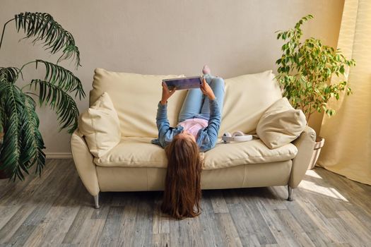 little girl lying upside down on sofa and play with tablet