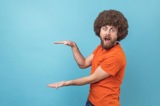 Portrait of amazed surprised man with Afro hairstyle presenting area for advertisement, looking at camera with big eyes and astonishment. Indoor studio shot isolated on blue background.
