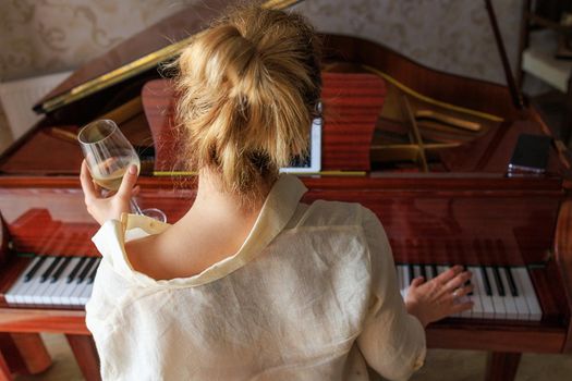 sad young blonde plays music on a retro piano, looks at notes in tablet. Sits back. The girl is holding a glass of wine in her hands. The concept of loneliness, depression, alcoholism in young people