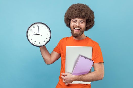 Portrait of satisfied man with Afro hairstyle wearing orange T-shirt holding paper notebook and laptop, showing big wall clock, finish work in time. Indoor studio shot isolated on blue background.