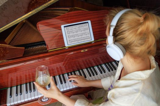 A young blonde girl, in white headphones, sits at the piano with her back. A woman holds a glass of white wine in her hands. The concept of loneliness, depression, alcoholism. Life style, close-up