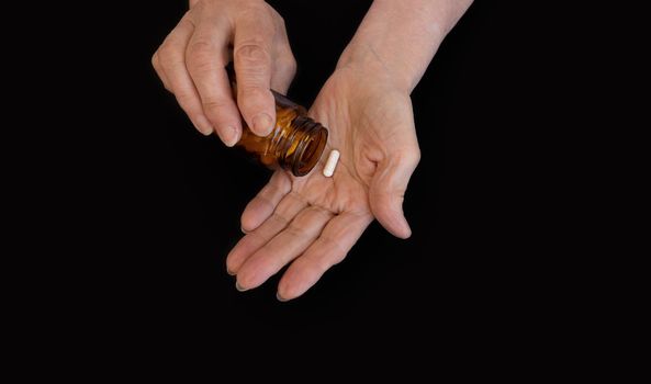 The hands of an elderly woman pour a pill from a jar into her palm. Black background, copy paste for your text. The concept of medicine, diseases of the elderly, prevention, taking vitamins