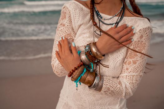 Model in boho style in a white long dress and silver jewelry on the beach. Her hair is braided, and there are many bracelets on her arms