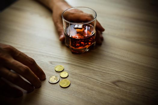 Fired man countsing his last money to drink expensive alcohol. Wrinkled alcoholic sits at brown wooden table with a glass of cognac with ice, on the table, empty wallet with coins