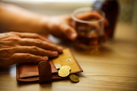 Fired man countsing his last money to drink expensive alcohol. Wrinkled alcoholic sits at brown wooden table with a glass of cognac with ice, on the table, empty wallet with coins