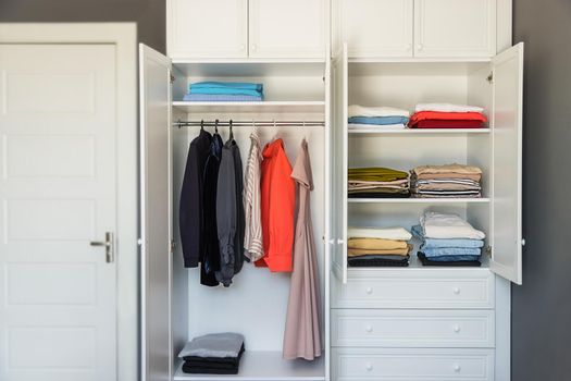Close-up of a white wardrobe with clothes. Modern dressing room, organization of space, reasonable consumption of things. Shirts, dresses and jackets are hung on hangers. T-shirts on the shelves