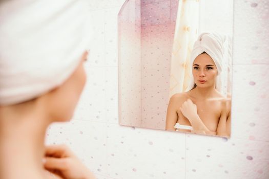 Portrait of young girl with towel on head in white bathroom looks and touches her face in the mirror and enjoys youth and hydration. Natural beauty, home care for problem skin.