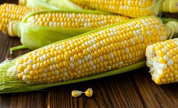 Fresh sweet corn with leaves on cobs on wooden table, closeup, top view. Fresh yellow corns ears with leaves. Ears of freshly harvested yellow sweet corn on wooden table.