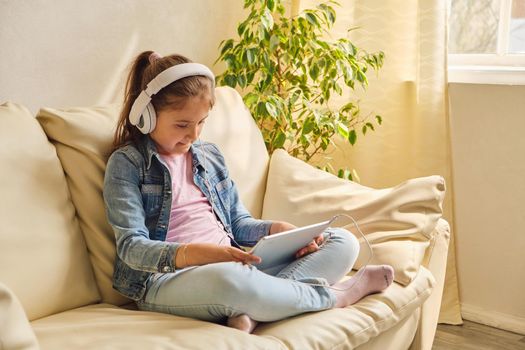 Cute little girl in casual clothes and headphones using a tablet and listening to music while sitting on a sofa in the room.