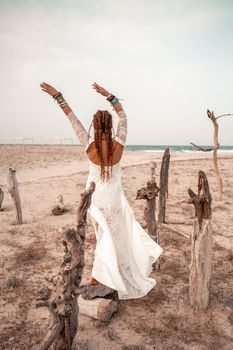 Model in boho style in a white long dress and silver jewelry on the beach. Her hair is braided, and there are many bracelets on her arms