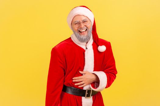 Portrait of elderly man with gray beard wearing santa claus costume holding his belly and laughing out loud, hearing funny story or joke. Indoor studio shot isolated on yellow background.