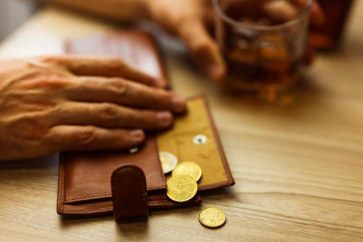 Fired man countsing his last money to drink expensive alcohol. Wrinkled alcoholic sits at brown wooden table with a glass of cognac with ice, on the table, empty wallet with coins