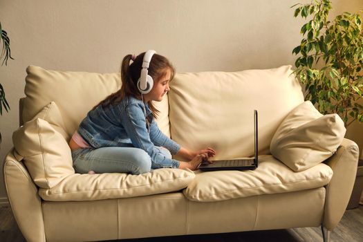 Online education. Kid girl with headphones looking video lesson teacher conference laptop sitting on the couch at home