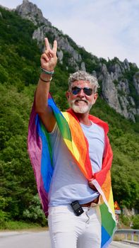 Gray-haired senior elderly diabetic man bisexuality with a beard and sunglasses with a rainbow LGBTQIA peace flag in mountains. Celebrates Pride Month, Rainbow Flag Day, gay parade. Vertical
