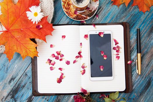 Autumn layout, notebook and pen, smartphone, cup of coffee, orange leaves, dry rose petals, knitted sweater. Cozy workplace, top view