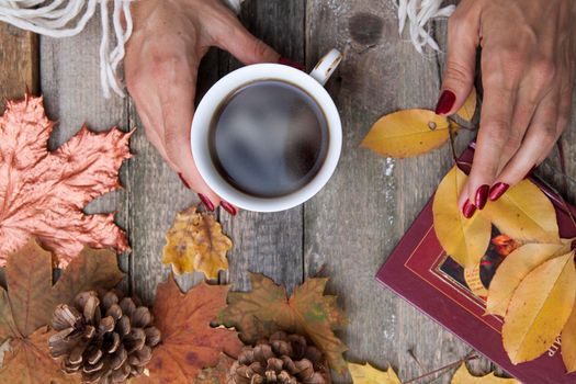 Top view cup of coffee in female hands on a wooden table background with autumn leaves, cones. Top view