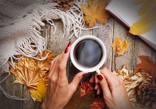Autumn layout: hot coffee in the hands of a girl, a book, golden autumn leaves, a knitted sweater, a plaid on a wooden table. Cozy autumn mood in October, November