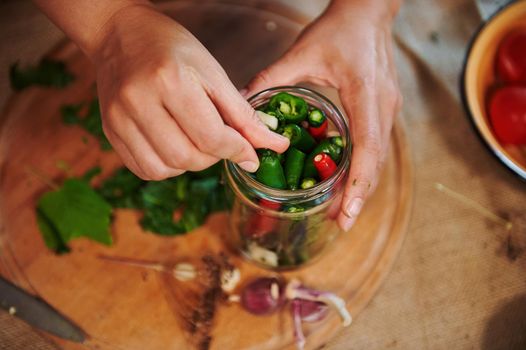 View from above of housewife hand adding fragrant culinary herbs and peeled fresh garlic cloves in freshly marinated red and green chili peppers. Close-up pickling, canning, marinating. Family recipe