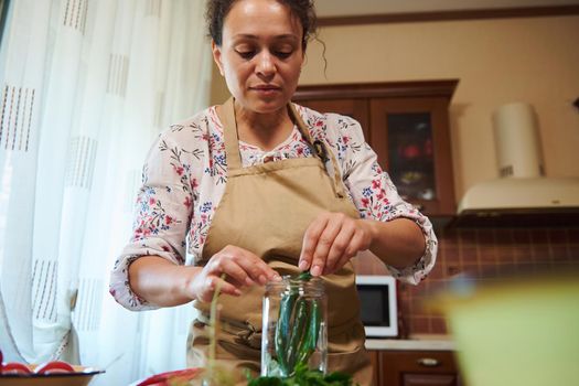 Pleasant housewife, chef in a beige apron, putting chili peppers into a sterilized glass can while canning in the home kitchen. Autumn seasonal canning for the winter. Homemade pickling