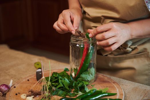 Close-up of the hands of a housewife putting umbrella dill and red green chili peppers while marinating spicy delicacies and making homemade preserves for the winter at home kitchen. Culinary. Canning