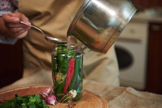 Close-up of a housewife pouring boiling brine or pickle over spicy chili peppers in glass while pickling seasonal vegetable at home kitchen. Harvest conservation. Marinating. Canning food for winter