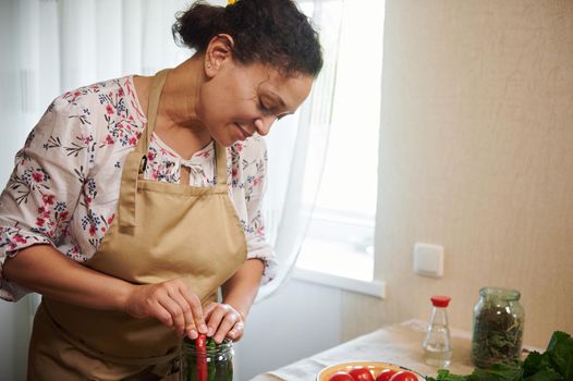 Portrait of a woman, housewife, filling glass jar for canning with an umbrella dill, organic chili peppers, fragrant culinary herbs while making homemade preserves and spicy delicacies for the winter