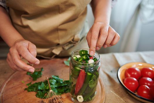 Cropped view of housewife's hand adding fragrant culinary herbs and peeled fresh garlic cloves in freshly marinated red and green chili peppers. Close-up pickling, canning, marinating. Family recipe