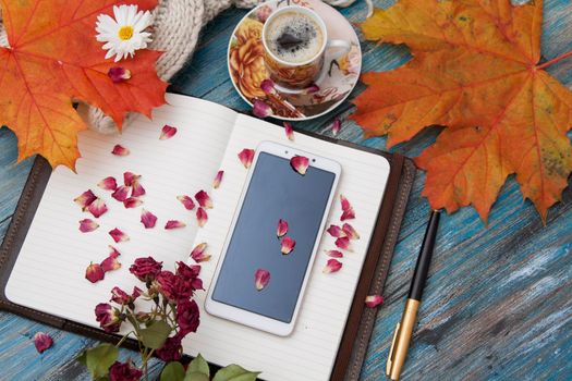 Autumn layout, a cup of coffee, yellow leaves, dry petals, a knitted sweater, a notebook, a smartphone. Cozy workplace, top view