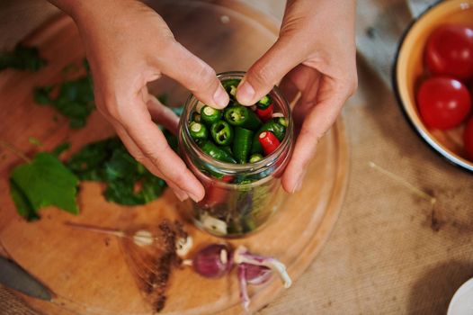 Details: Hands of a housewife, putting ingredients and stacking chili peppers into a sterilized glass jar while canning in the home kitchen. Autumn seasonal canning for the winter. Homemade pickling