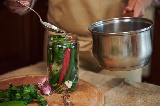 Close-up. Housewife pours boiling marinade over chili peppers in jars. Chopped dill leaves and fresh garlic laying out on a wooden board. Pickling seasonal vegetable at home. Harvest conservation.