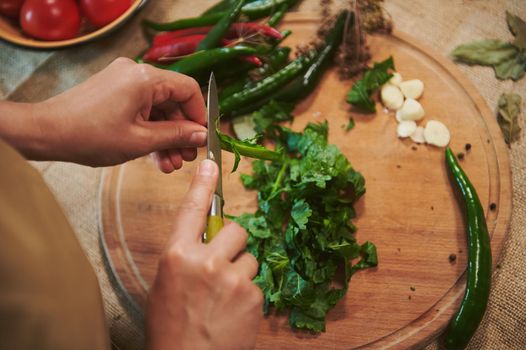 Top view of the hands of a housewife chopping fresh fragrant culinary herbs on a wooden cutting board while preparing a pickle marinade for pickling chili peppers in the home kitchen. homemade food