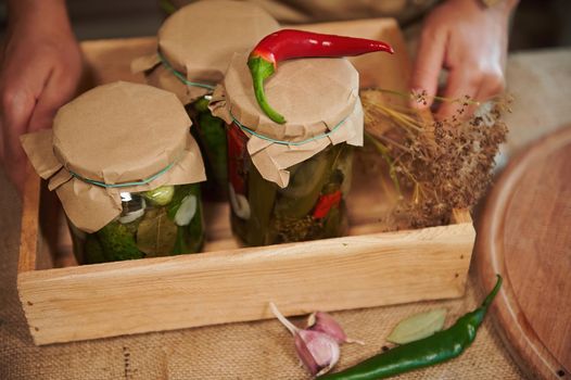 Cropped view of a wooden crate with jars of delicious homemade tasty pickled cucumbers and marinated chili peppers. Fresh ingredients on a table with a linen tablecloth