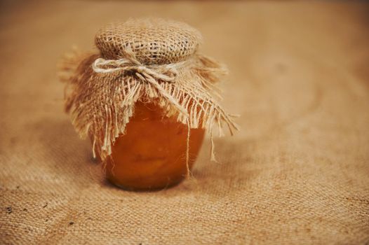 Still life. Jar of homemade peach jam with a burlap on the lid, on the table with linen tablecloth. Canned goods. Confiture canned according traditional family recipe. Copy space for advertising text