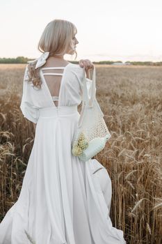 A blonde woman in a long white dress walks in a wheat field. The concept of a wedding and walking in nature.