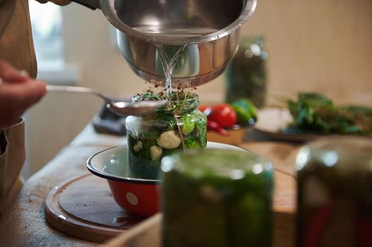 Close-up of pouring boiling marinade or brine over cucumbers, from a saucepan in jar, while pickling organic vegetable harvest at home kitchen. Jars with canned food upside down on blurred foreground