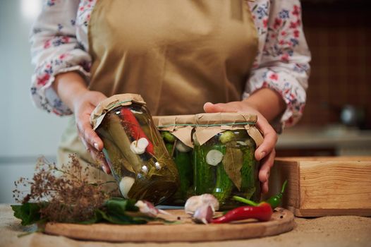 Details: hands of a housewife demonstrate to the camera homemade canned food, pickles, pickled chili peppers and marinated cucumbers, on a wooden board with fresh garlic cloves, umbrella dill and herbs